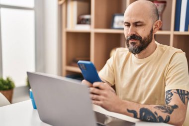 Evdeki masada oturan genç kel adam dizüstü bilgisayar ve akıllı telefon kullanıyor.