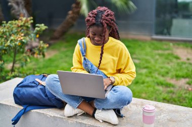 African american woman student using laptop sitting on bench at campus park