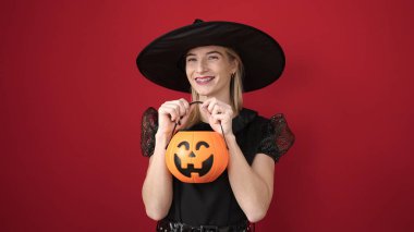 Young blonde woman wearing witch costume holding pumpkin basket over isolated red background