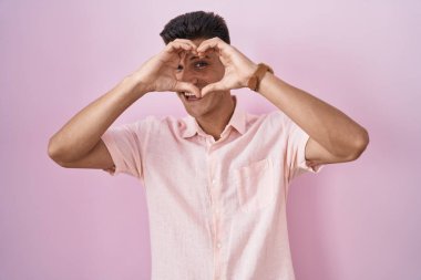 Young hispanic man standing over pink background doing heart shape with hand and fingers smiling looking through sign 