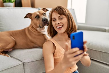 Kafkasyalı genç bir kadın akıllı telefonun yanında evde köpekle oturmuş selfie çekiyor.