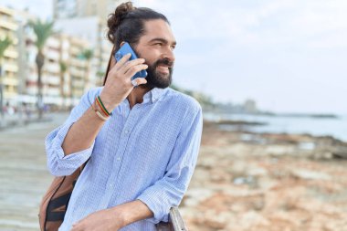 Young hispanic man musician talking on smartphone holding guitar case at seaside