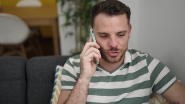 Young caucasian man speaking on the phone sitting on the sofa at home