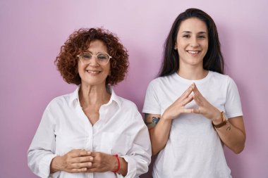 İspanyol anne ve kızı pembe arka plan elleri üzerine gündelik beyaz tişört giyiyorlar ve mutlu ve rahat bir şekilde gülümsemeyi diliyorlar. Başarı ve iyimserlik 