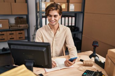 Young caucasian man ecommerce business worker using smartphone writing on notebook at office