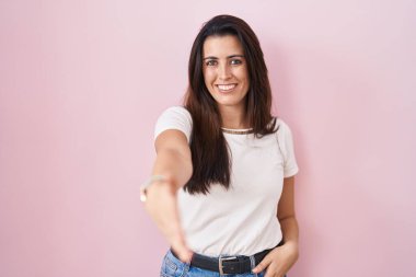 Pembe arka planda duran esmer genç bir kadın selamlama ve hoş geldin demek için dostça bir şekilde tokalaşıyor. başarılı iş. 