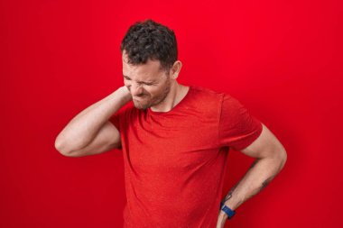 Young hispanic man standing over red background suffering of neck ache injury, touching neck with hand, muscular pain 