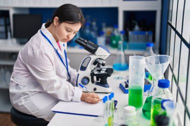 Down sendromlu genç bir kadın. Laboratuvarda mikroskop kullanarak belgelere yazı yazıyor.