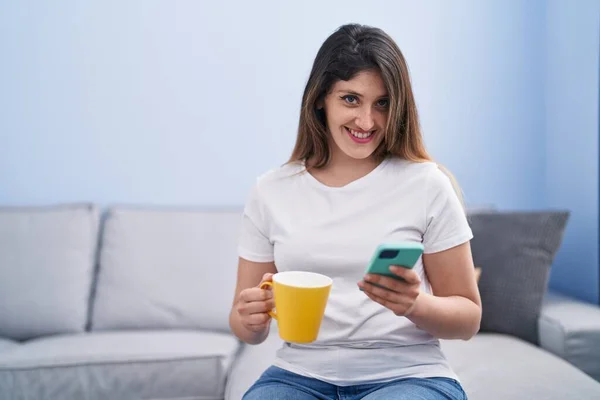 若いヒスパニック系の女性がコーヒーを飲み 自宅でスマートフォンを使用して — ストック写真