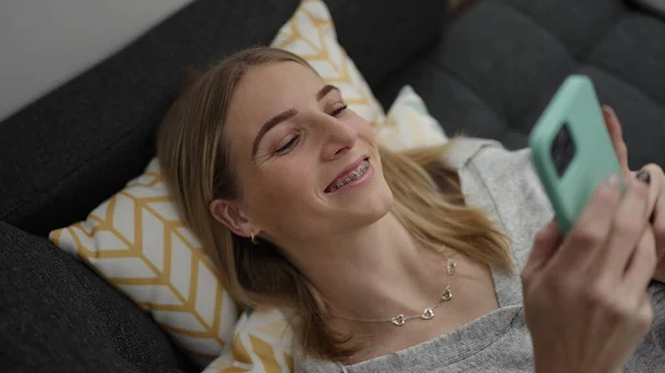 Young Blonde Woman Using Smartphone Lying Sofa Home — Stok fotoğraf