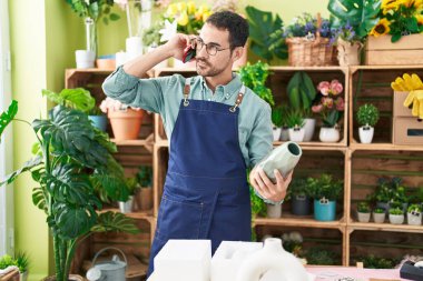 Young hispanic man florist talking on smartphone holding plant pot at flower shop
