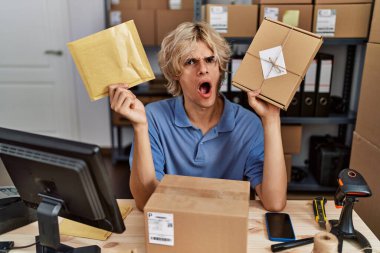 Küçük işletme şirketinde çalışan genç adam şok olmuş suratıyla paketleri tutuyor, şüpheci ve alaycı görünüyor, ağzı açık kalınca şaşırıyor. 