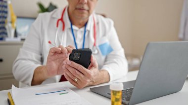 Gri saçlı, orta yaşlı bir adam klinikte çalışan akıllı telefonu kullanıyor.
