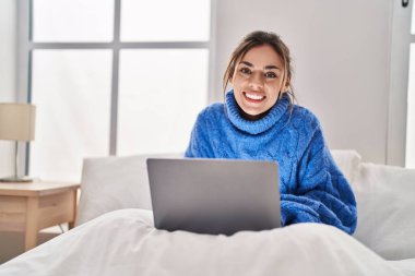 Young beautiful hispanic woman using laptop sitting on bed at bedroom