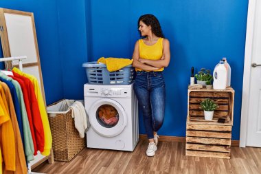 Genç Latin kadın gülümsüyor. Kollarını kavuşturup çamaşırhanede duruyor.