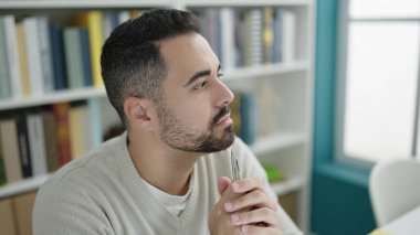 Young hispanic man student sitting on table with doubt expression at library university