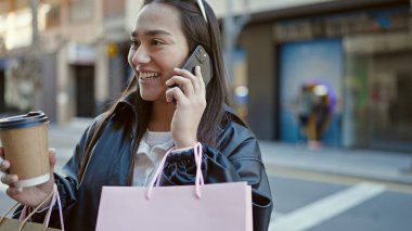 Güzel İspanyol bir kadın akıllı telefondan konuşuyor. Elinde alışveriş poşetleri ve sokakta kahve var.