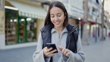 Genç, güzel, İspanyol bir kadın sokakta akıllı telefon kullanarak gülümsüyor.