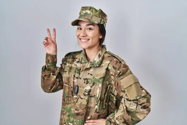 Young south asian woman wearing camouflage army uniform smiling looking to the camera showing fingers doing victory sign. number two.