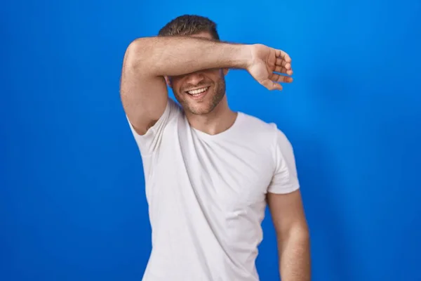 青い背景の上に立つ若い白人男性は 陽気で面白い腕で目を覆います 視覚障害の概念 — ストック写真