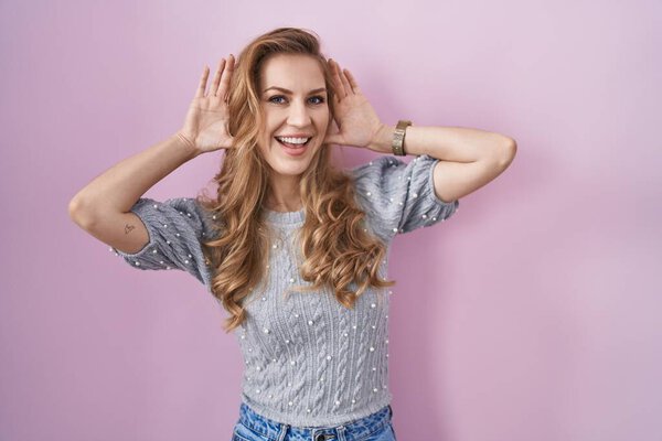 Beautiful blonde woman standing over pink background smiling cheerful playing peek a boo with hands showing face. surprised and exited 