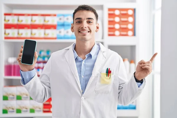 Красивый Латиноамериканец Работающий Аптеке Показывает Экран Смартфона Улыбающийся Счастливо Указывая — стоковое фото