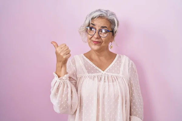 Middle age woman with grey hair standing over pink background smiling with happy face looking and pointing to the side with thumb up.