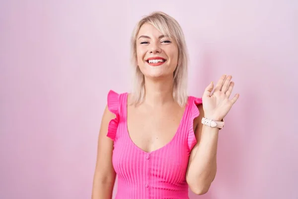 若いです白人女性がピンクの背景に立って手を振っ挨拶幸せと笑顔 優しい歓迎ジェスチャー — ストック写真
