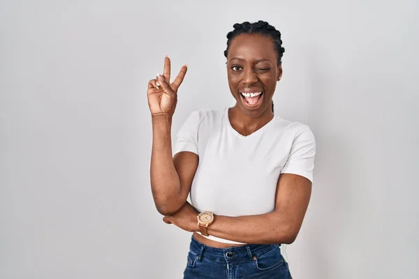 Beautiful black woman standing over isolated background smiling with happy face winking at the camera doing victory sign with fingers. number two.