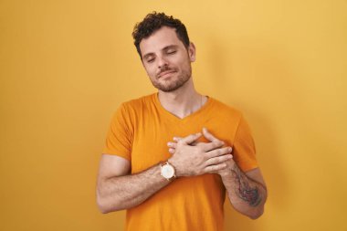 Young hispanic man standing over yellow background smiling with hands on chest with closed eyes and grateful gesture on face. health concept. 