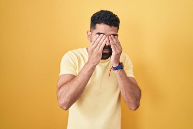 Hispanic man with beard standing over yellow background rubbing eyes for fatigue and headache, sleepy and tired expression. vision problem 