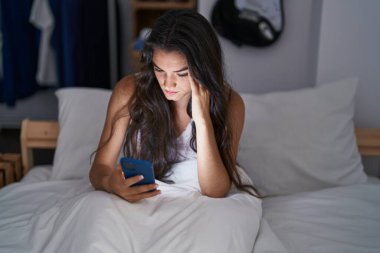 Young hispanic woman using smartphone stressed on bed at bedroom