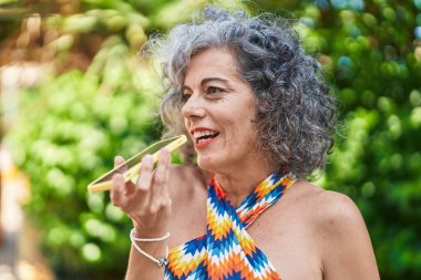 Orta yaşlı, kır saçlı, gülümseyen, kendine güvenen bir kadın parkta akıllı telefondan konuşuyor.