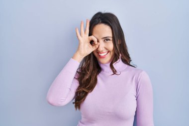 Genç esmer kadın mavi arka planda duruyor. El hareketleriyle gülümsüyor, gözleriyle mutlu yüzüyle parmaklarının arasından bakıyor.. 