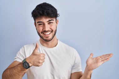 Hispanic man with beard standing over white background showing palm hand and doing ok gesture with thumbs up, smiling happy and cheerful 