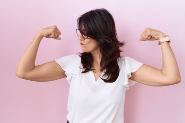 Orta yaşlı İspanyol kadın, sıradan beyaz tişört ve gözlük takıyor. Kollarında gururla gülümseyen kaslar var. fitness konsepti. 