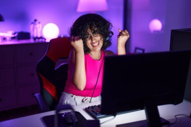 Young middle eastern woman streamer playing video game with winner expression at gaming room