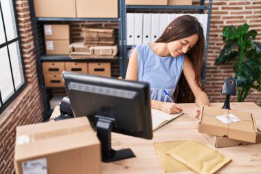 Young beautiful hispanic woman ecommerce business worker using computer writing on notebook at office