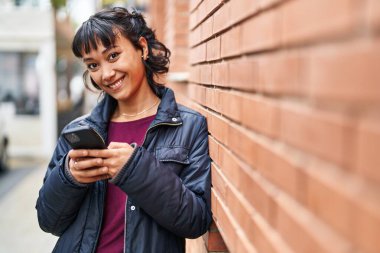 Genç, güzel, İspanyol bir kadın sokakta akıllı telefon kullanarak gülümsüyor.