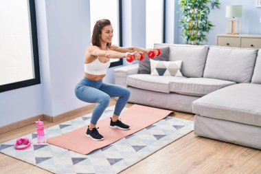 Young beautiful hispanic woman training leg exercise using dumbbells at home