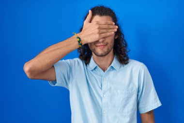 Young hispanic man standing over blue background smiling and laughing with hand on face covering eyes for surprise. blind concept. 