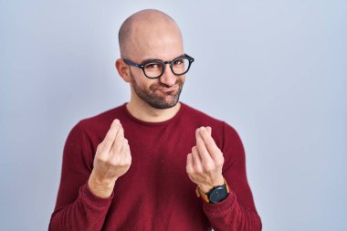 Beyaz arka plan üzerinde duran sakallı genç bir adam gözlük takıyor. Elleriyle para hareketi yapıyor. Maaş ödemesi istiyor. 