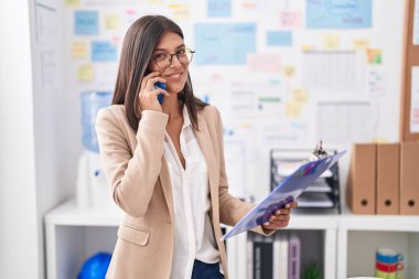 Young beautiful hispanic woman business worker talking on smartphone holding clipboard at office