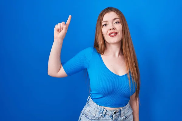 自信と幸せ笑顔ながら 青色の背景に立っている赤毛の女性が表示され 指のナンバーワンで指摘します — ストック写真