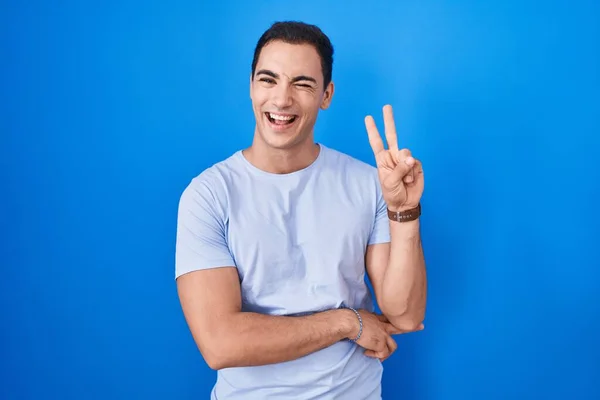 青い背景の上に立っている若いヒスパニック系の男は カメラで喜んで顔をウィンクしながら笑顔で指で勝利サインをしています — ストック写真