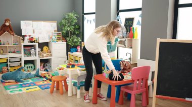 Young blonde woman preschool teacher picking up toys at kindergarten