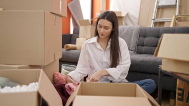 Young beautiful hispanic woman unpacking cardboard box at new home