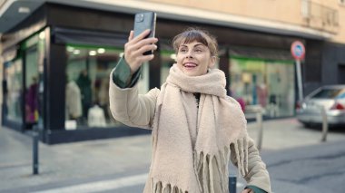 Beyaz bir kadın sokakta akıllı telefonuyla selfie çekiyor.