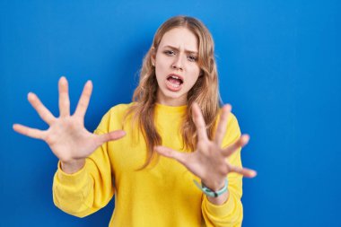 Young caucasian woman standing over blue background afraid and terrified with fear expression stop gesture with hands, shouting in shock. panic concept. 