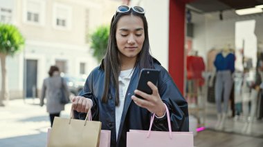 Genç, güzel, İspanyol bir kadın akıllı telefon kullanıyor. Elinde alışveriş torbaları var.